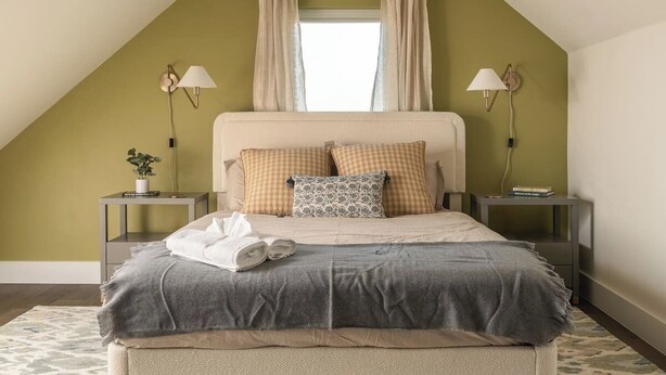 A bedroom with a sloped ceiling, a bed with a grey blanket, two bedside tables with lamps, and a window with curtains. - Pool, Mini-golf, Sauna, Movie, Gym | Sleep 18 (Austin)