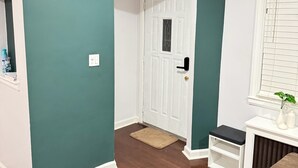 Um corredor com uma porta branca, um relógio na parede, um degrau pequeno e uma janela com persianas.