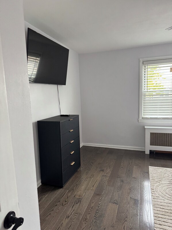 A room with a flat-screen TV mounted on the wall, a black storage unit with drawers, a window with blinds, and a wooden floor.
