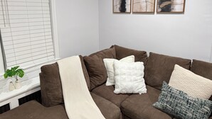 Uma sala de estar com um sofá marrom, almofadas brancas e estampadas, uma janela com persianas e três quadros abstratos na parede.