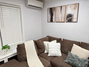 Ein Wohnzimmer mit einem braunen Sofa, weißen und gemusterten Kissen, einem Fenster mit Jalousien und drei abstrakten Bildern an der Wand.