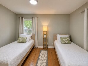 Ein Schlafzimmer mit zwei Betten, einem Nachttisch mit Lampe, einem Fenster mit Vorhängen und Holzboden.