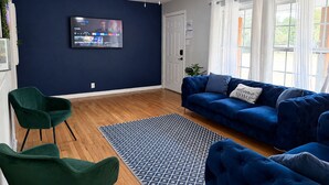 A living room with a blue sofa, green chairs, a patterned rug, and a television on the wall. - Family-Friendly 3BR – 1 Mi to Six Flags, AT&T Stadium & Globe Life Field (Arlington)
