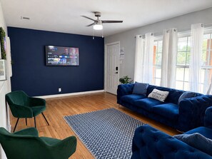 Ein Wohnzimmer mit einem blauen Sofa, grünen Sesseln, einem gemusterten Teppich und einem Fernseher an der Wand.