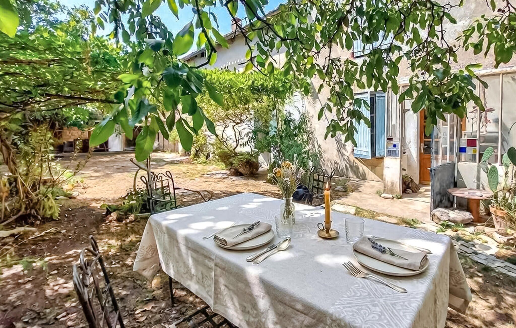 Un espace extérieur aménagé pour prendre les repas, avec une table dressée pour deux personnes, entouré de verdure et avec une maison en arrière-plan.