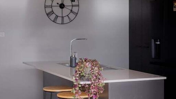 A modern kitchen with a central island, bar stools, a wall-mounted clock, and a hanging light fixture.