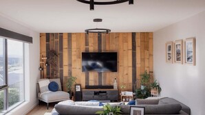 A modern living room with a large flat-screen TV mounted on a wooden wall, a grey sofa, a small dining table with chairs, and a large window providing a view of the outdoors.