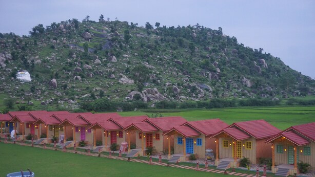 A row of traditional-style buildings with red roofs, set against a backdrop of a green hill with scattered rocks. - Vatika Green Resort & Restaurant  (Devgadh Bariya)