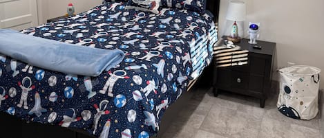 A bedroom with a bed, a ceiling fan, an air conditioner, a nightstand, a lamp, and a wall with space-themed decorations.