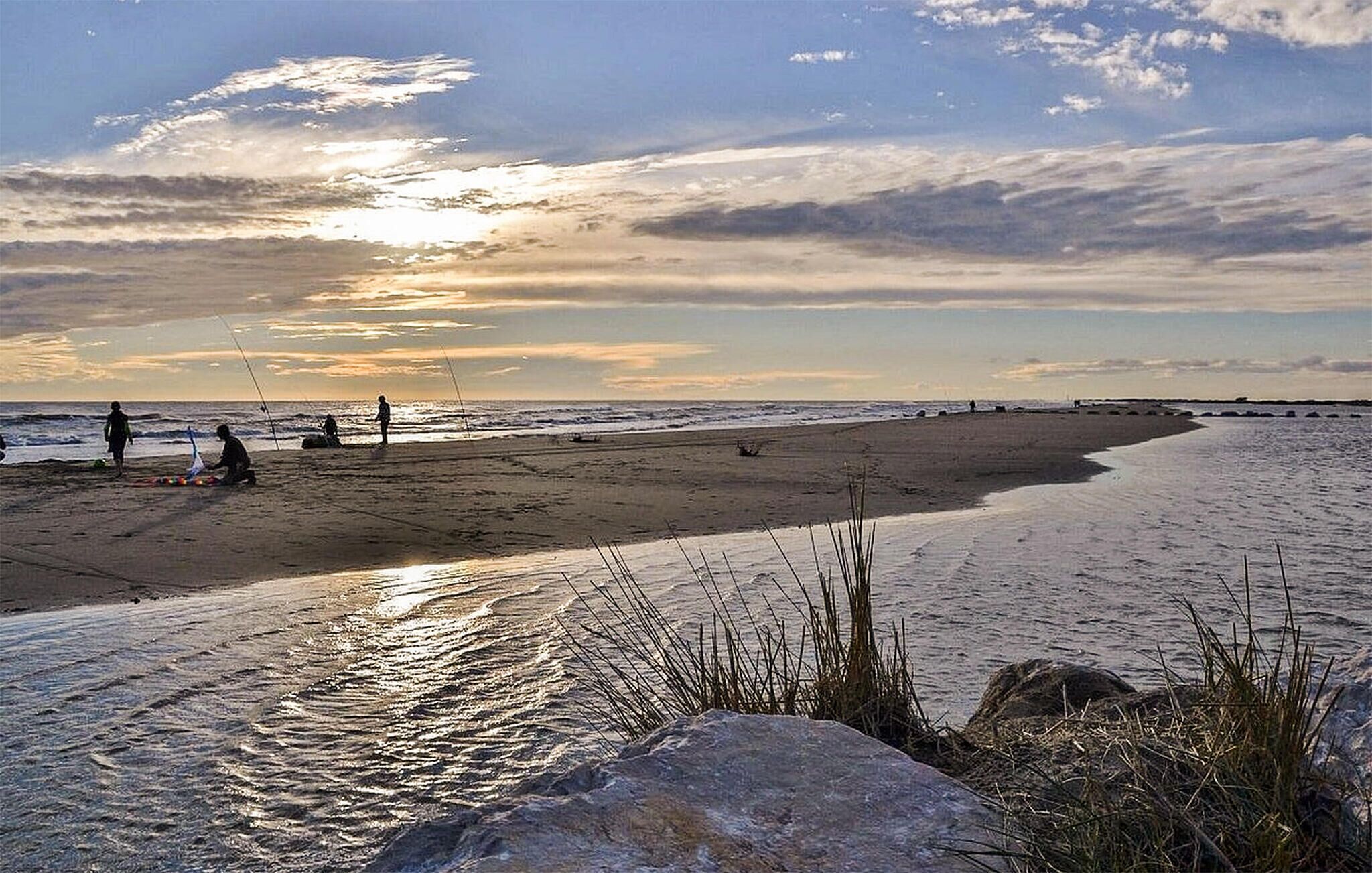 Una playa con gente pescando y caminando, un mar tranquilo y un cielo al atardecer.