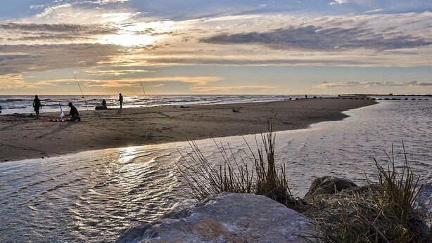 Een strand met mensen die aan het vissen zijn, kalm water en een zonsondergang.