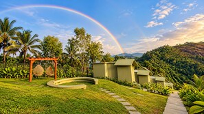 A lush garden with a curved pathway, a wooden pergola with hanging chairs, and a row of green-roofed buildings. - Grandlivet Resort (Vythiri)
