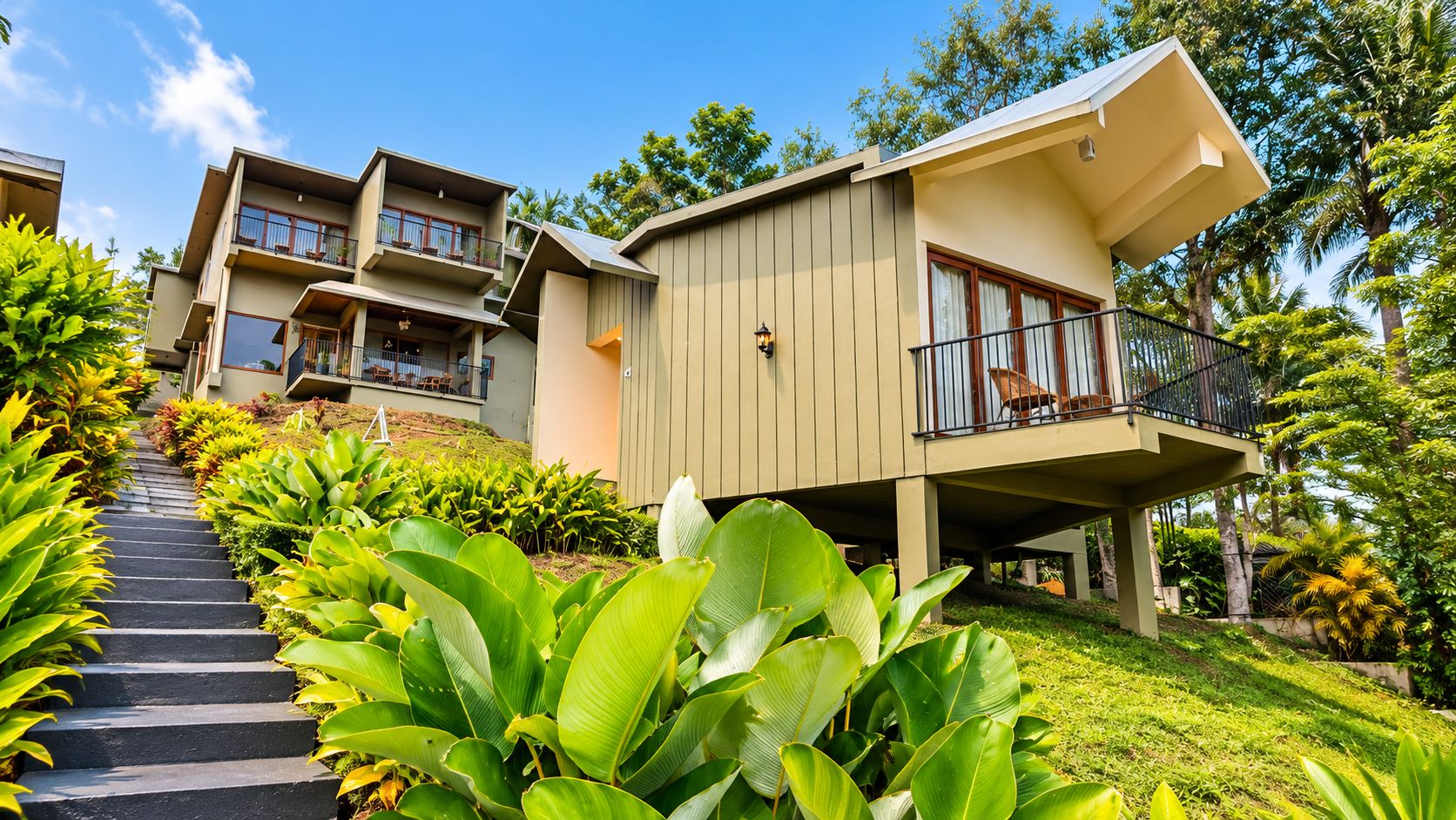 A modern multi-story building with balconies, surrounded by lush greenery and a clear sky.