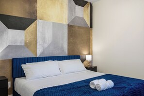 A hotel room with a bed, pillows, a blue headboard, a nightstand with a lamp, and a wall with geometric patterns. - The Encore - Rooftop Hot Tub/Game Room/Sleeps 12 (Nashville)