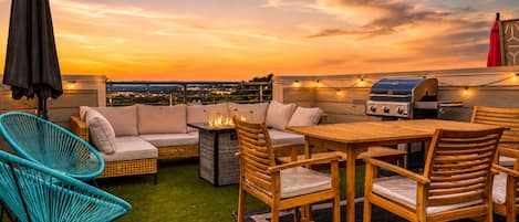 A rooftop patio with a grill, seating area, and a view of the cityscape at sunset.