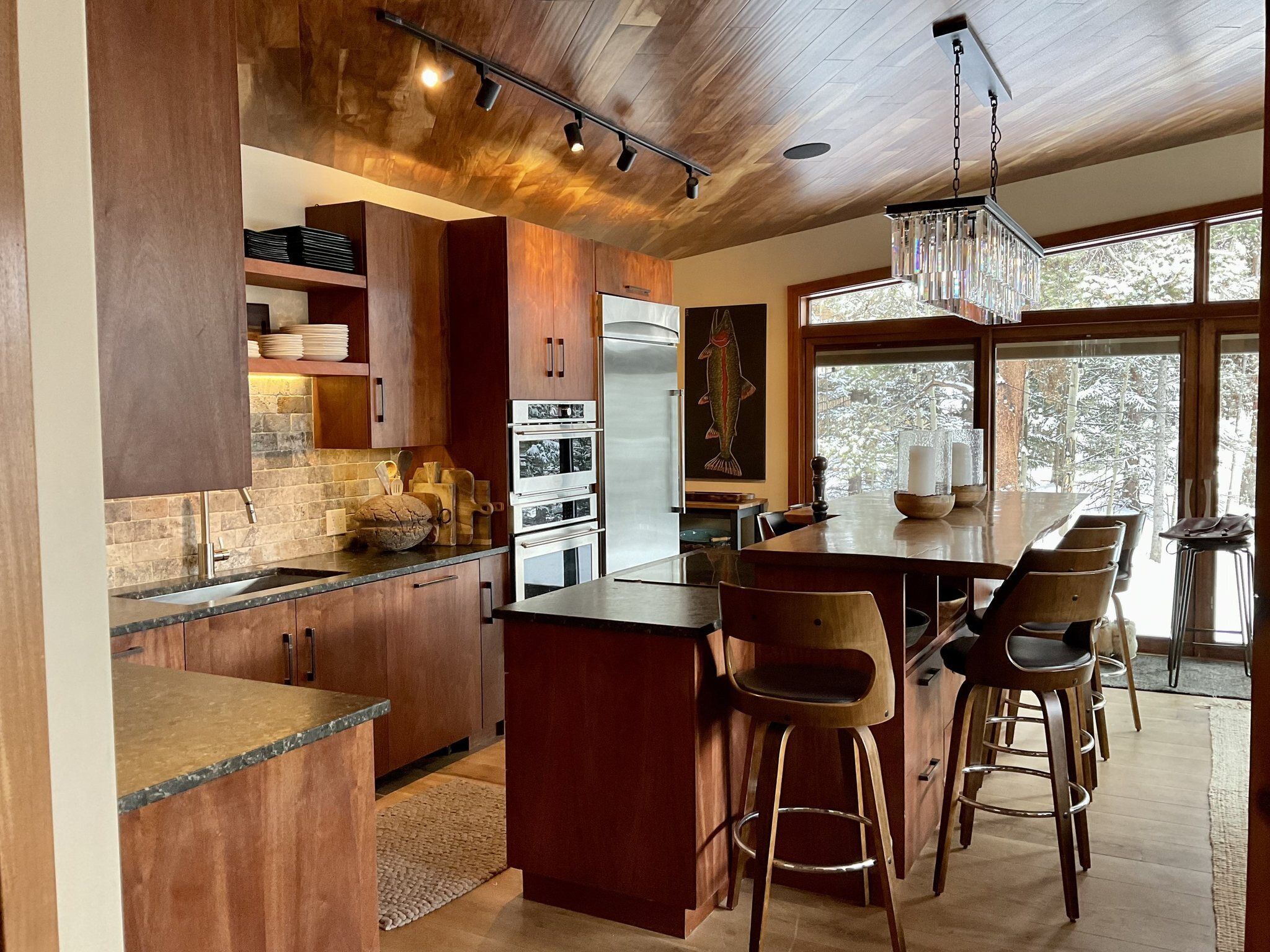 A modern kitchen with wooden cabinets, a central island, bar stools, and a large window providing a view of the outdoors.