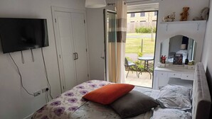 Une chambre à coucher avec un lit, un téléviseur fixé au mur, une fenêtre donnant sur l’extérieur et une petite table avec une lampe.