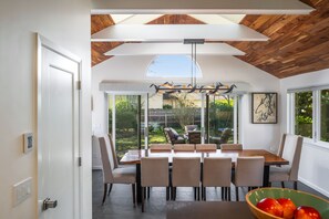 A dining area with a large table and chairs, a view of the outdoors, and a ceiling with wooden beams. - Magnolia Oasis (Seattle)