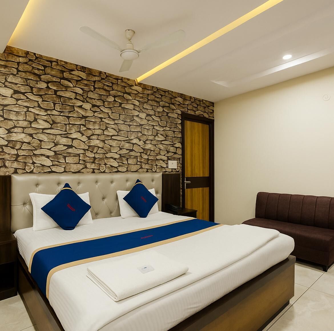 Habitación de hotel con pared de piedra, cama con detalles azules, sofá marrón y ventilador de techo.