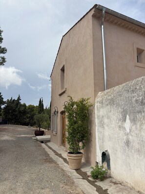 A building with a beige facade, a visible window, and a door. There is a large planter with a tree next to the building. - Maison de Garrigue Estanh – A charming estate with a pool near Narbonne (Bages)