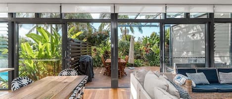 A modern outdoor living space with a wooden table and chairs, a sofa, and a glass wall offering a view of lush greenery.
