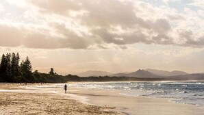 A beach with a person walking, trees, and mountains in the distance. - Kiah 9 Belongil Beach (Byron)