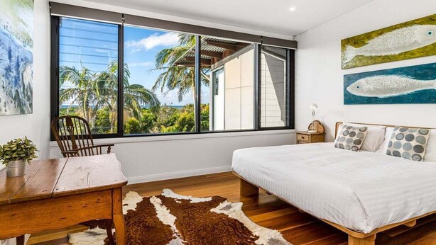 A bedroom with a large window, a ceiling fan, a bed with white bedding, and a cowhide rug. - Kiah 9 Belongil Beach (Byron)