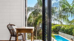 A room with a wooden table and chair, a large window with a view of a swimming pool and palm trees. - Kiah 9 Belongil Beach (Byron)