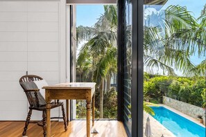 Ein Zimmer mit Holztisch und Stuhl, ein großes Fenster mit Blick auf einen Swimmingpool und Palmen.
