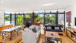 A modern living room with a large glass wall, a dining area with a wooden table and chairs, and a seating area with sofas and a coffee table. - Kiah 9 Belongil Beach (Byron)