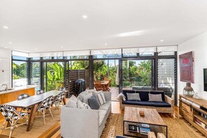 Ein modernes Wohnzimmer mit einer großen Glasscheibe, einem Essbereich mit Holztisch und -stühlen sowie einer Sitzecke mit Sofas und einem Couchtisch.