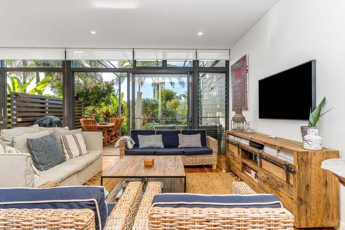 A modern living room with a sofa, coffee table, and wicker furniture, large windows, and a flat-screen TV.