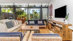A modern living room with a sofa, coffee table, and wicker furniture, large windows, and a flat-screen TV. - Kiah 9 Belongil Beach (Byron)