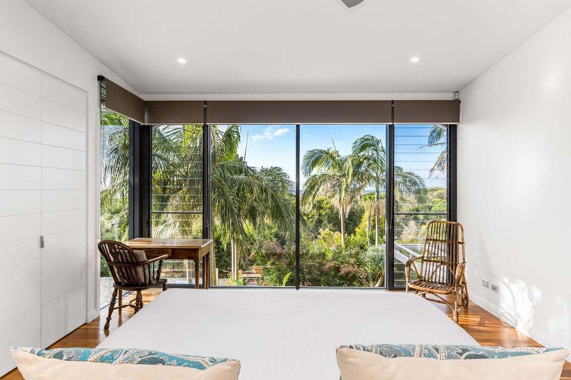 A bedroom with a large bed, two chairs, a dining table, and a view of a tropical garden through floor-to-ceiling windows.