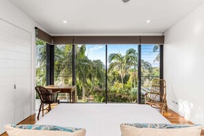 Ein Schlafzimmer mit einem großen Bett, zwei Stühlen, einem Esstisch und Blick auf einen tropischen Garten durch raumhohe Fenster.
