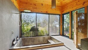Un baño moderno con una bañera grande, techo de madera y amplios ventanales con vistas a zonas verdes.