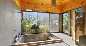 A modern bathroom with a large bathtub, wooden ceiling, and large windows offering a view of greenery.