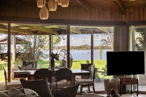 A room with a dining table and chairs, a television, and large windows overlooking a body of water.