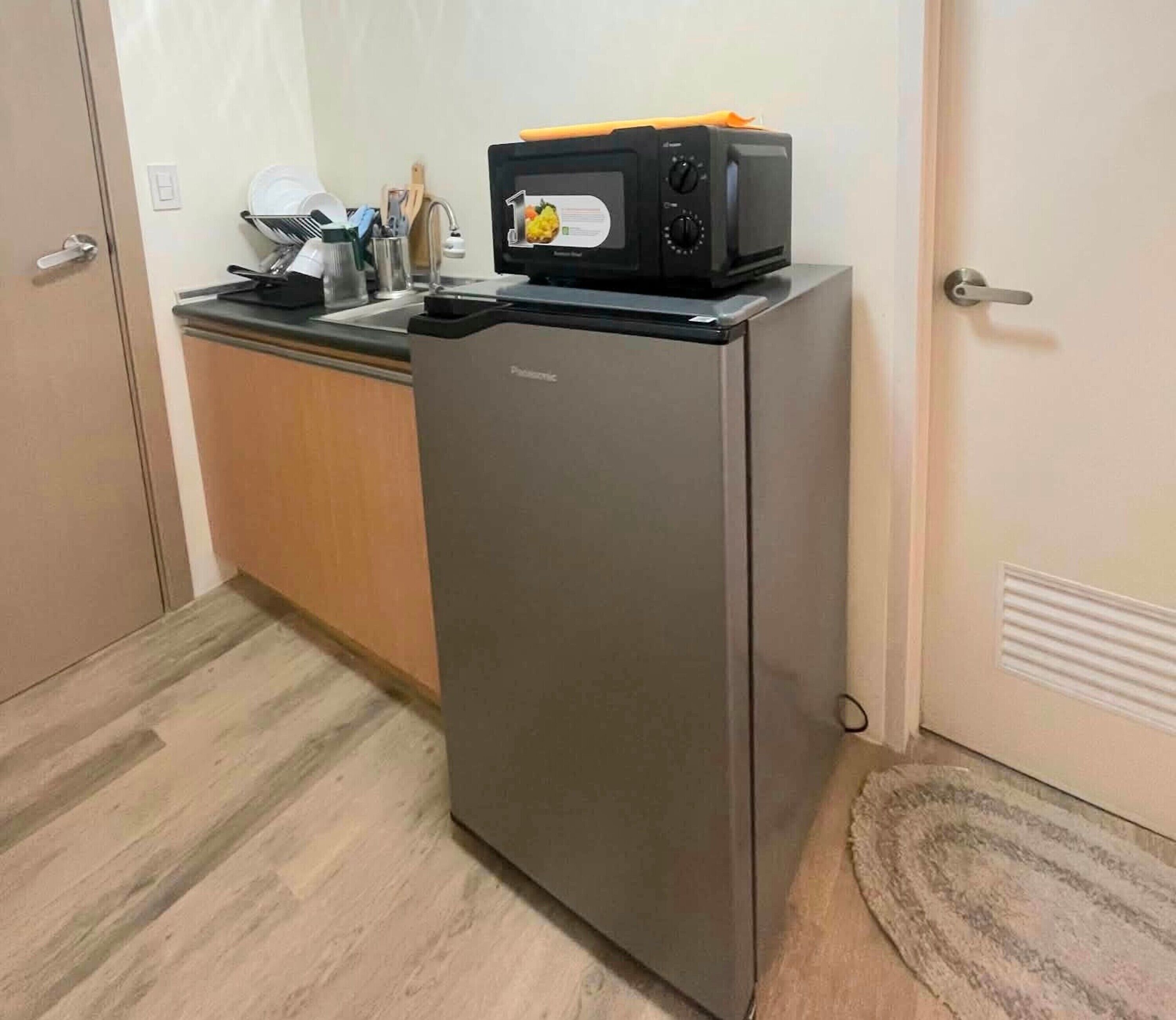 A compact kitchen with a stainless steel refrigerator, a microwave on top, and a sink with dishes.