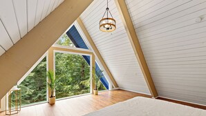 Et soveværelse med skrå vægge, en seng, en hængende lampe og udsigt til grøn natur.