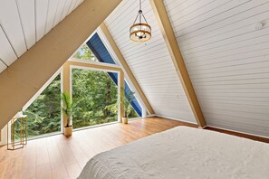 A bedroom with a sloped ceiling, a bed, a hanging light fixture, and a view of greenery outside.