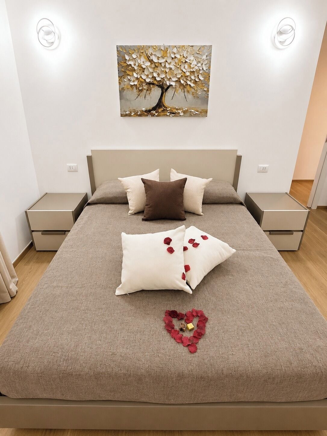 A neatly made bed with a beige bedspread, two pillows, and a heart-shaped arrangement of rose petals.