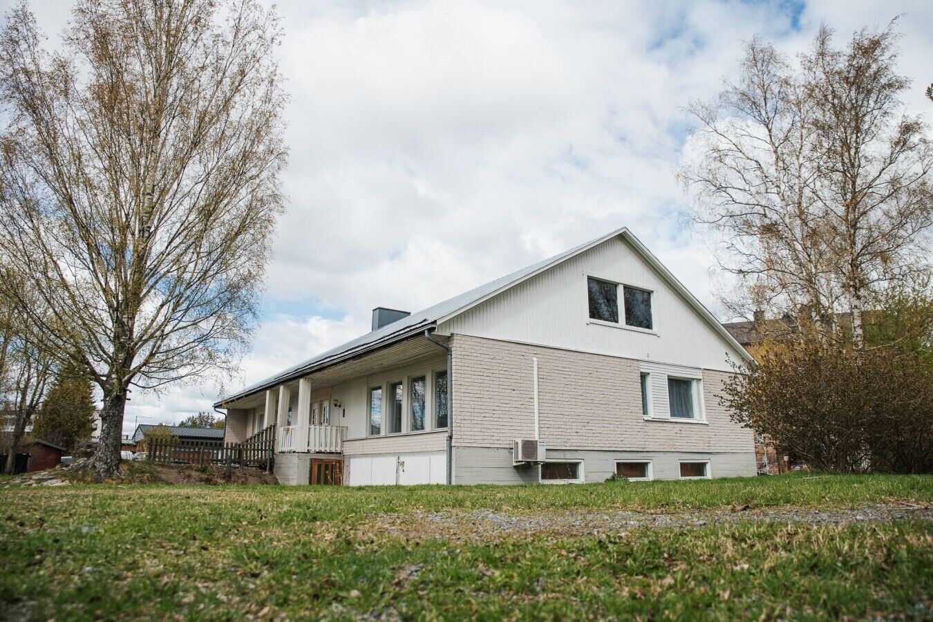 Kaksikerroksinen talo, jossa on kuisti, savupiippu ja suuri puu etualalla.