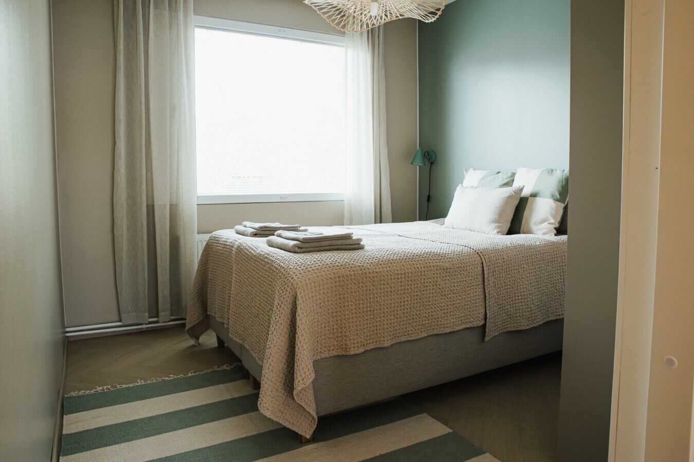 Un dormitorio con cama, una ventana con cortinas, una lámpara de noche y una alfombra a rayas.
