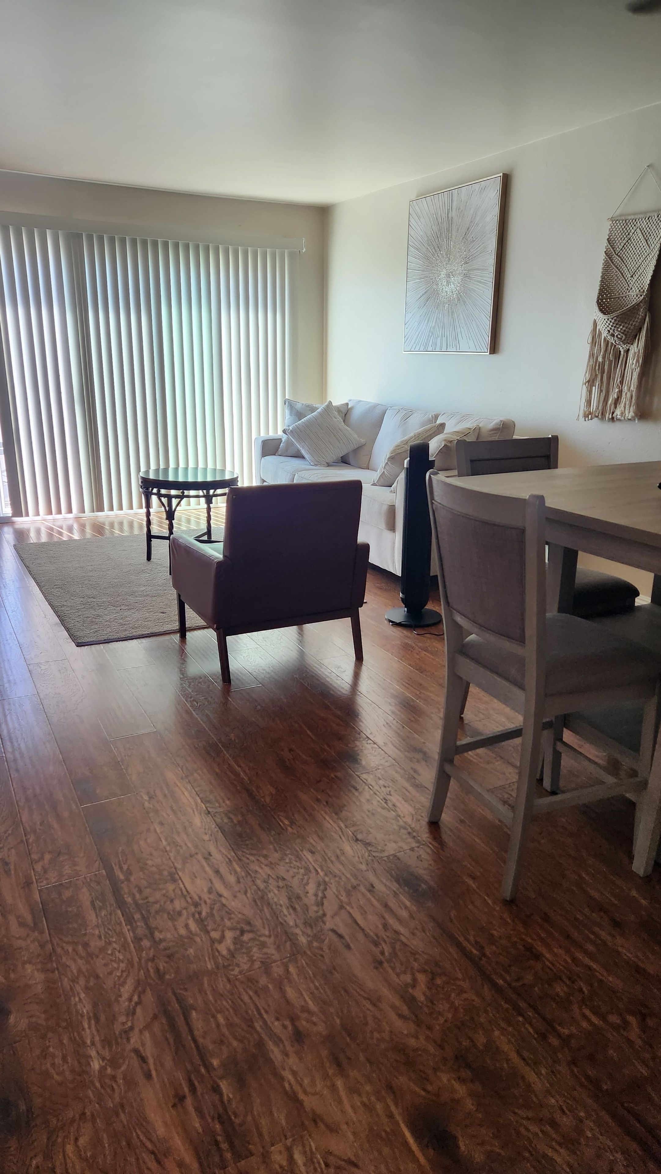 A living room with a sofa, a coffee table, a dining table with chairs, and a large window with blinds.