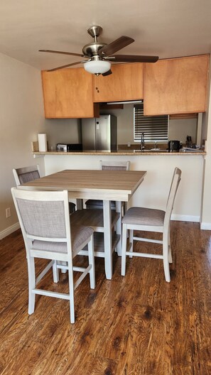 Een eetruimte met een houten tafel en stoelen, een plafondventilator, en een keuken met kastjes en apparatuur.