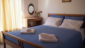 A neatly made bed with blue bedding, two folded towels, a wooden headboard, and a nightstand with a mirror and flowers. - Amalia House Garden (Pefki)