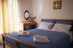 Ein ordentlich bezogenes Bett mit blauer Bettwäsche, zwei gefaltete Handtücher, ein hölzernes Kopfteil und ein Nachttisch mit Spiegel und Blumen.