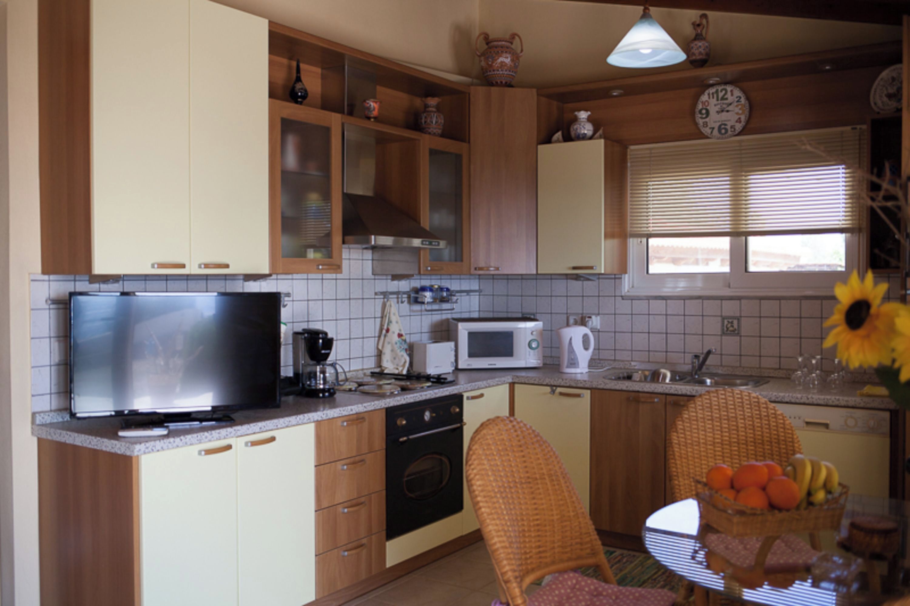 A kitchen with wooden cabinets, a microwave, a coffee maker, and a dining area with a wicker chair and a fruit basket.