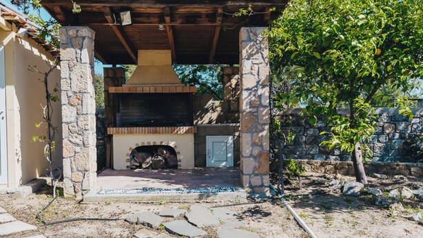 An outdoor barbecue area with a stone chimney, a covered seating area, and a stone pathway leading to the entrance. - Amalia House Garden (Pefki)